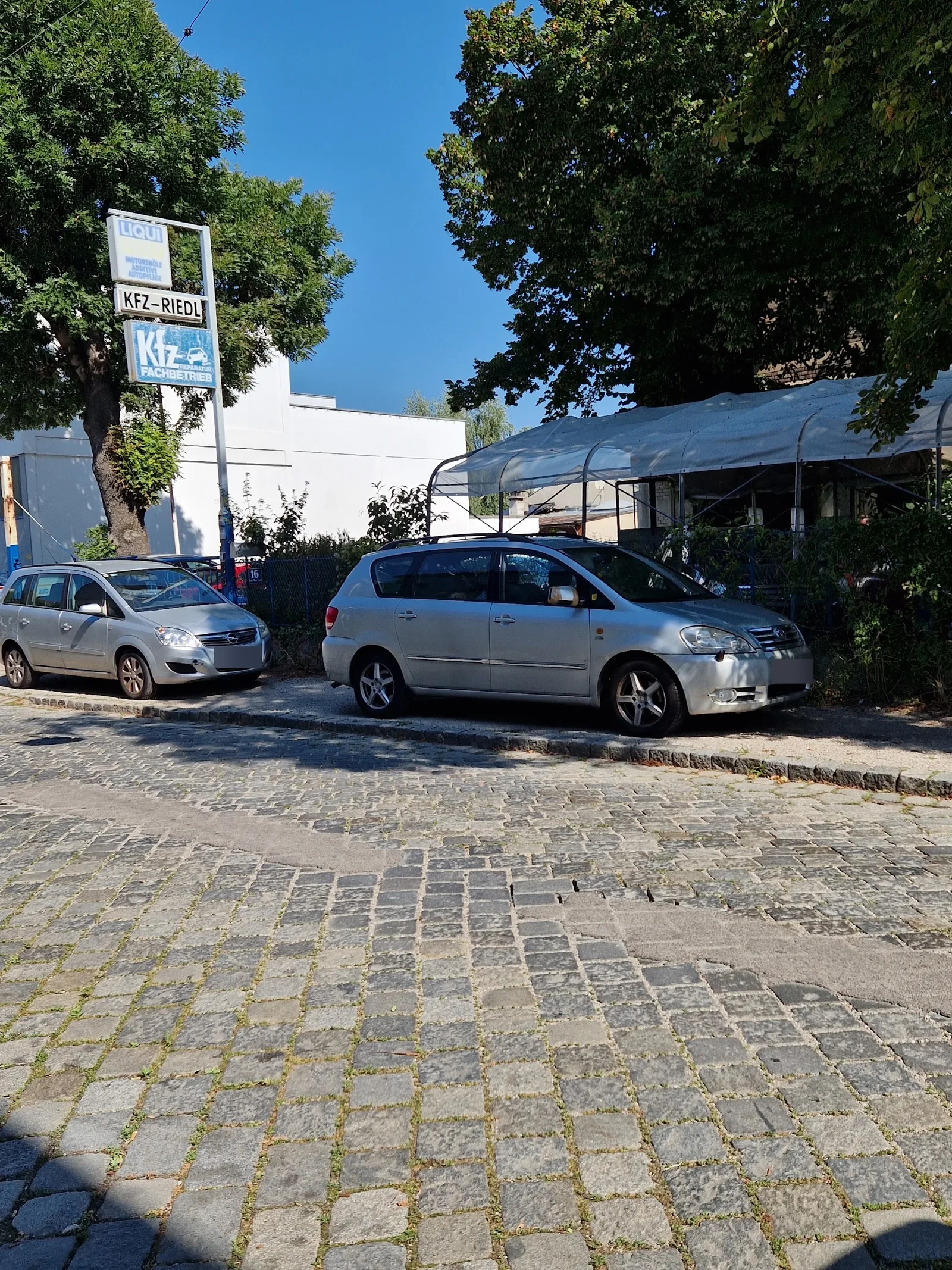 Schildermast mit den Tafeln KFZ-RIEDL und Kfz Fachbetrieb, davor geparkte Autos