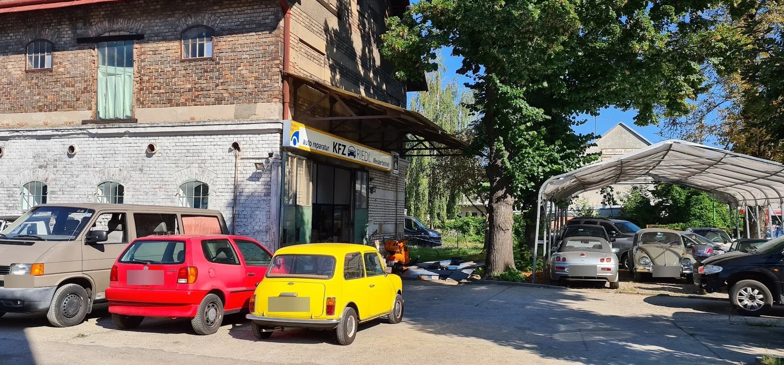 Gelber Mini, roter Polo und ein VW Käfer vor einer Werkstatthalle mit dem Schild KFZ RIEDL Meisterbetrieb